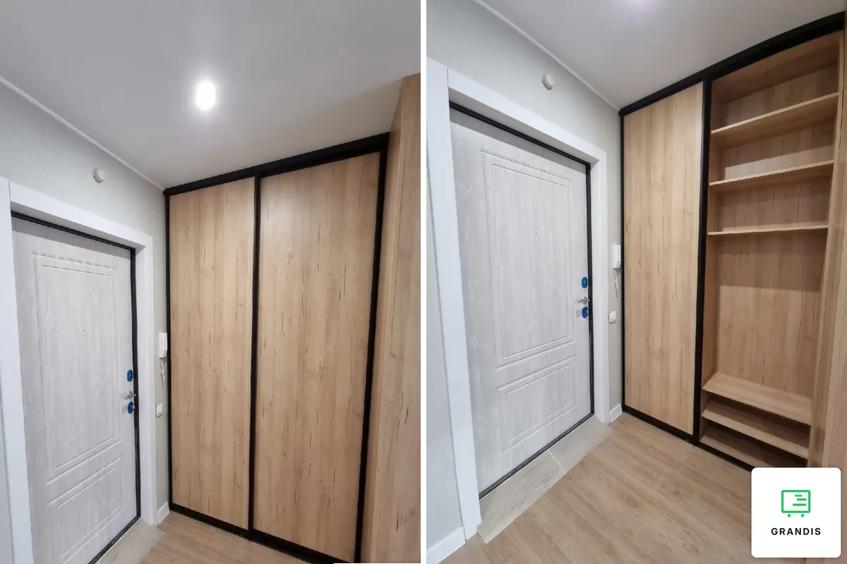 Oak-effect sliding wardrobe with a black frame closing a niche by the front door; next to it the same wardrobe with a door slid aside and shelves inside
